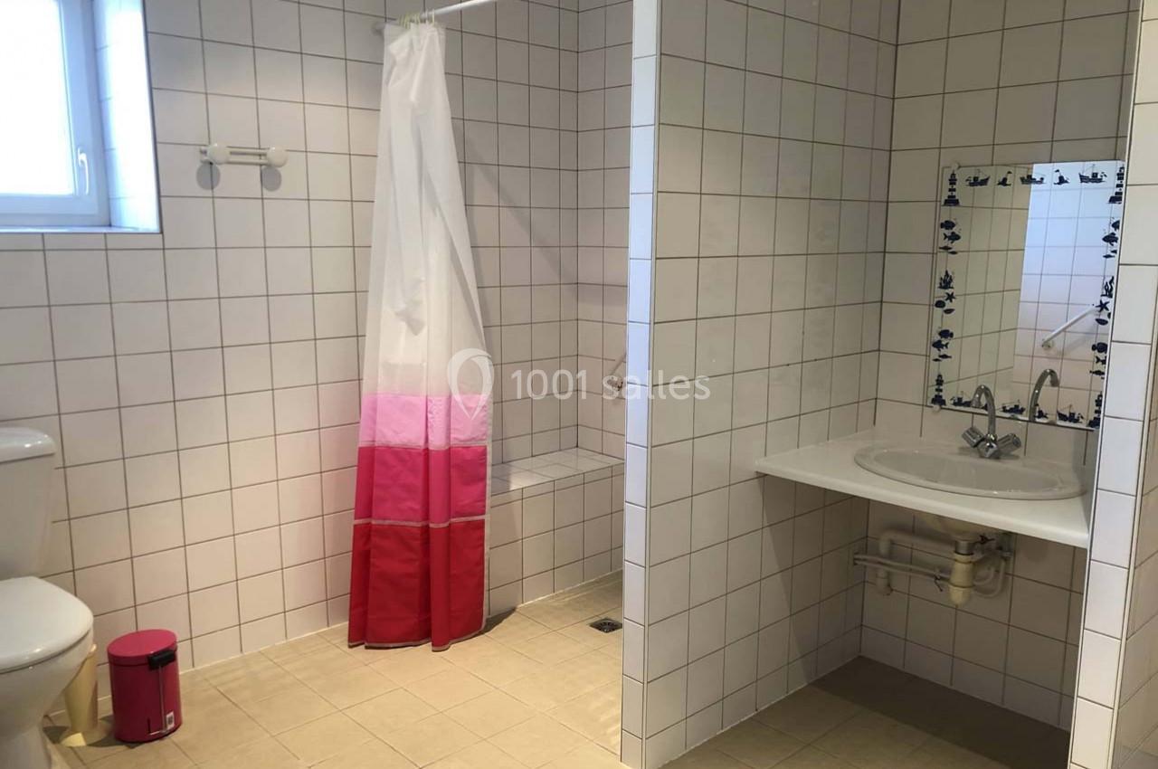 Salle de bain avec douche ouverte, rideau coloré, lavabo, miroir encadré et toilettes sur un carrelage blanc.