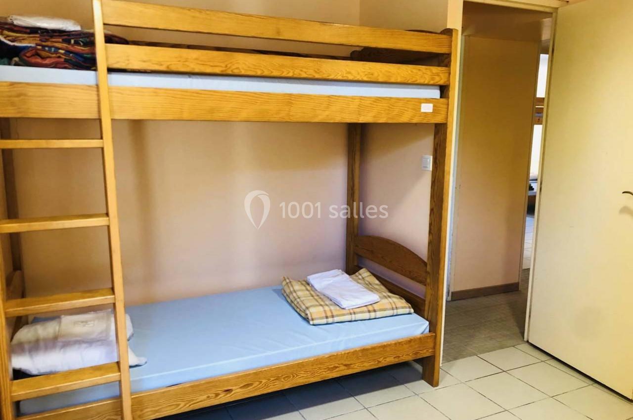 Lit superposé en bois dans une chambre simple avec draps pliés sur le matelas inférieur et porte ouverte sur un couloir.