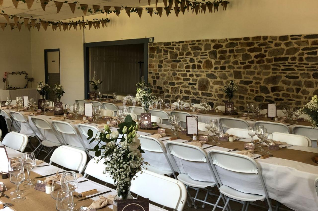 Salle décorée pour un événement avec tables alignées, nappes blanches, fleurs et guirlandes suspendues.
