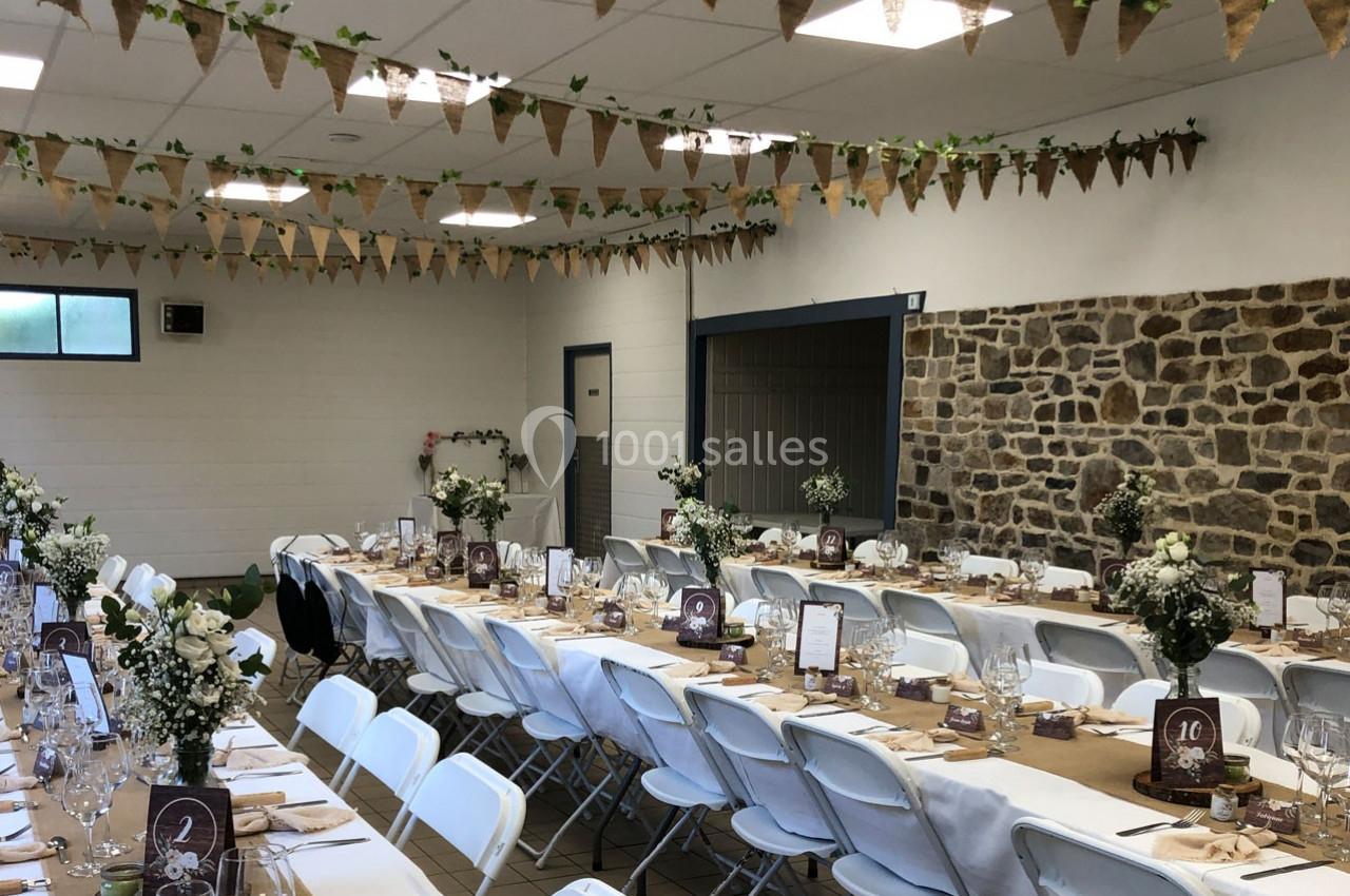 Salle décorée pour un événement, avec longues tables dressées, chaises blanches et guirlandes suspendues.
