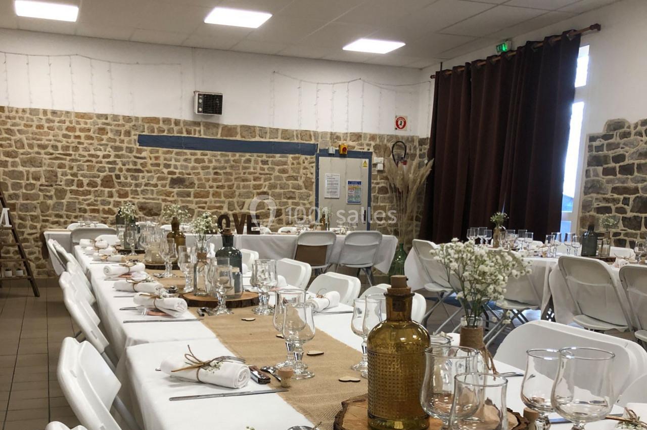 Salle décorée pour un événement avec des tables alignées, nappes blanches, décorations rustiques et murs en pierre.