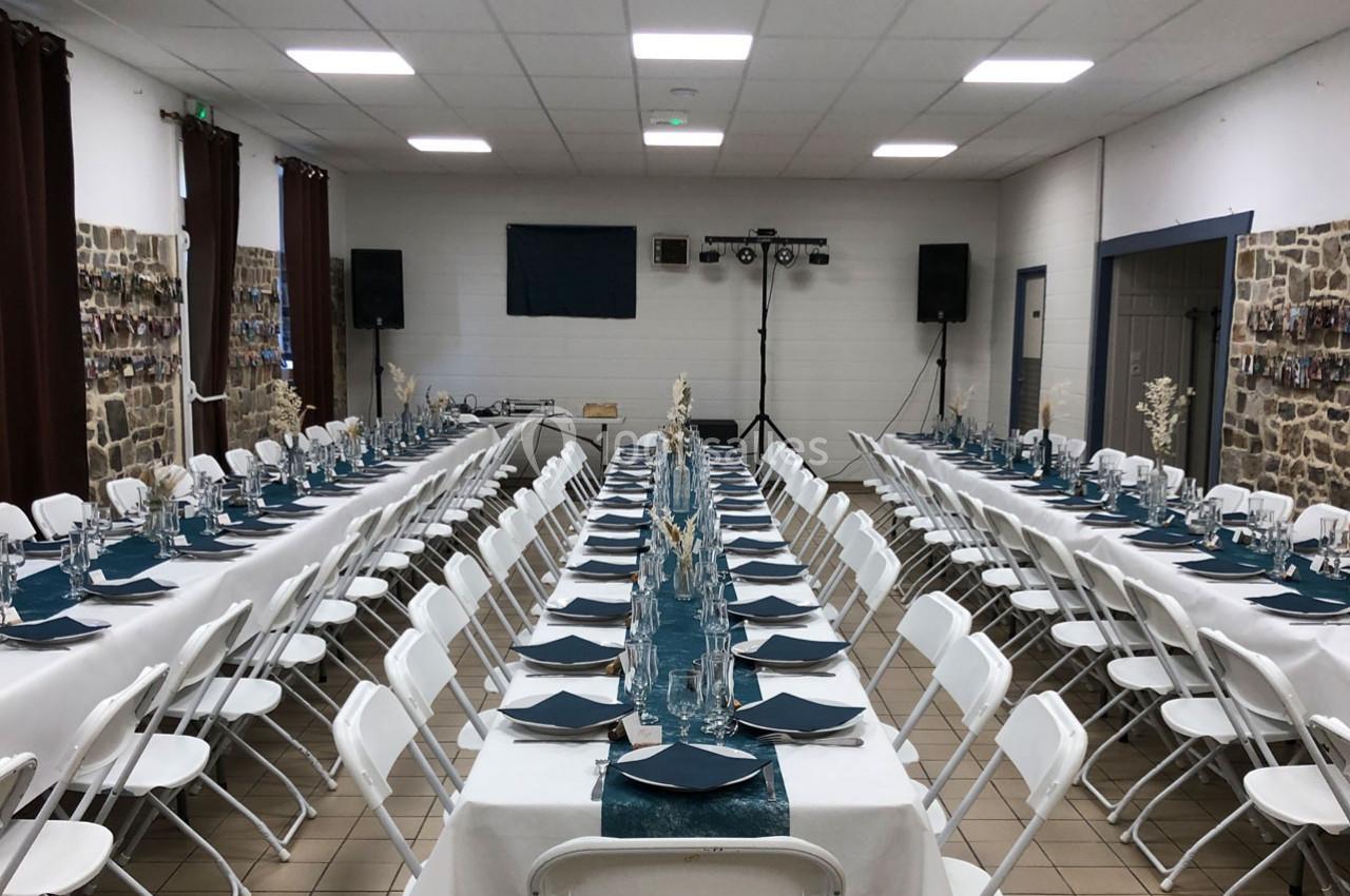 Salle de réception aménagée avec deux longues tables dressées, nappes blanches et décorations bleues, prête pour un…