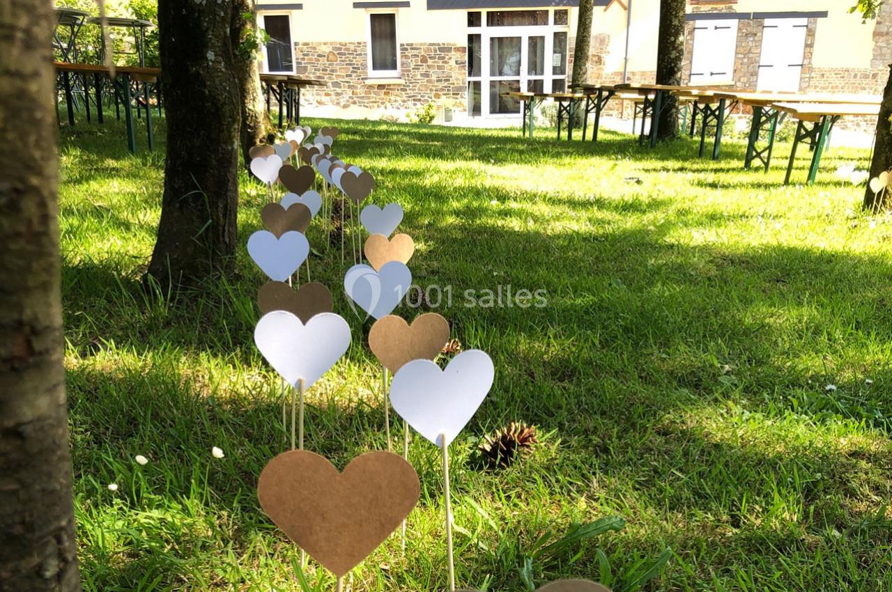 Cœurs en papier blanc et marron plantés sur des tiges dans une pelouse, avec des tables et un bâtiment en arrière-plan.