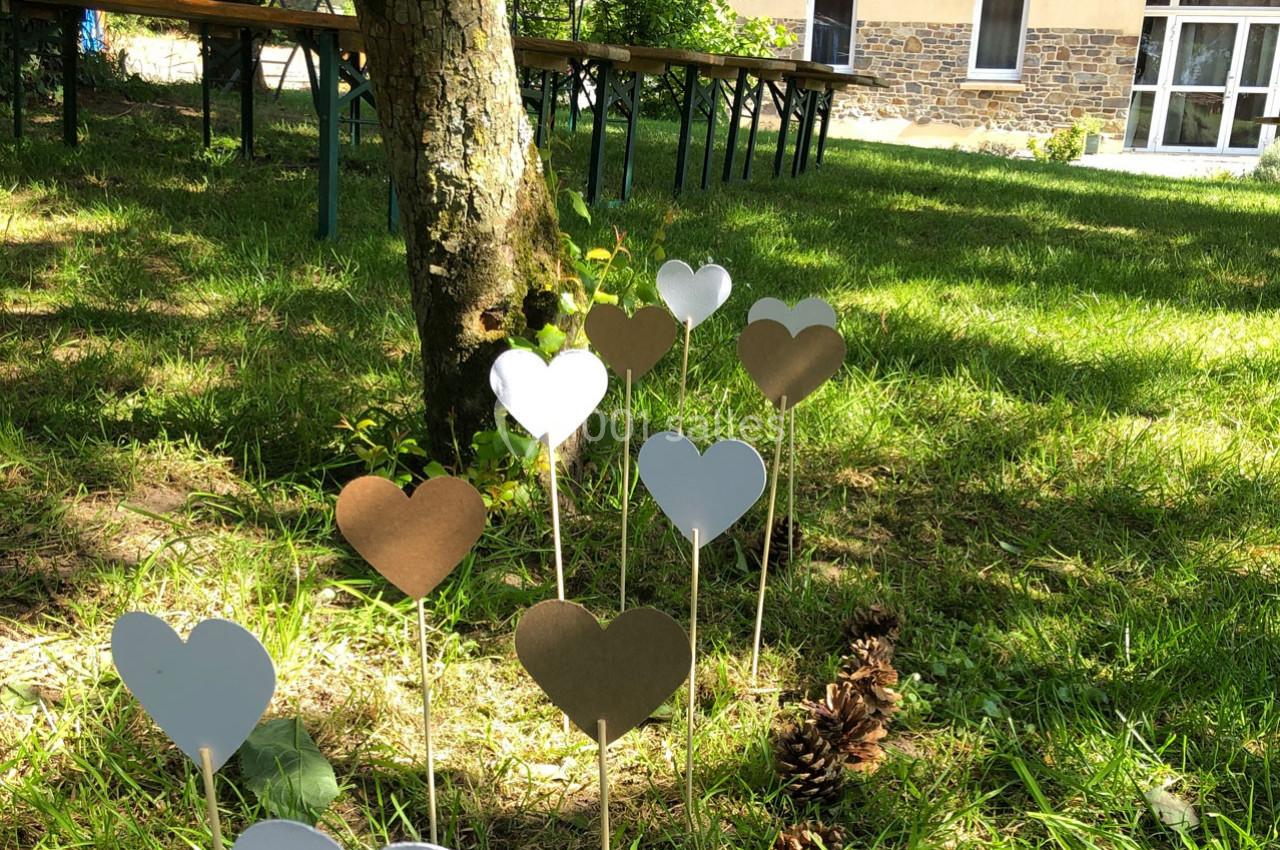 Cœurs en papier blanc et doré plantés dans l'herbe sous un arbre, avec des tables et un bâtiment en arrière-plan.