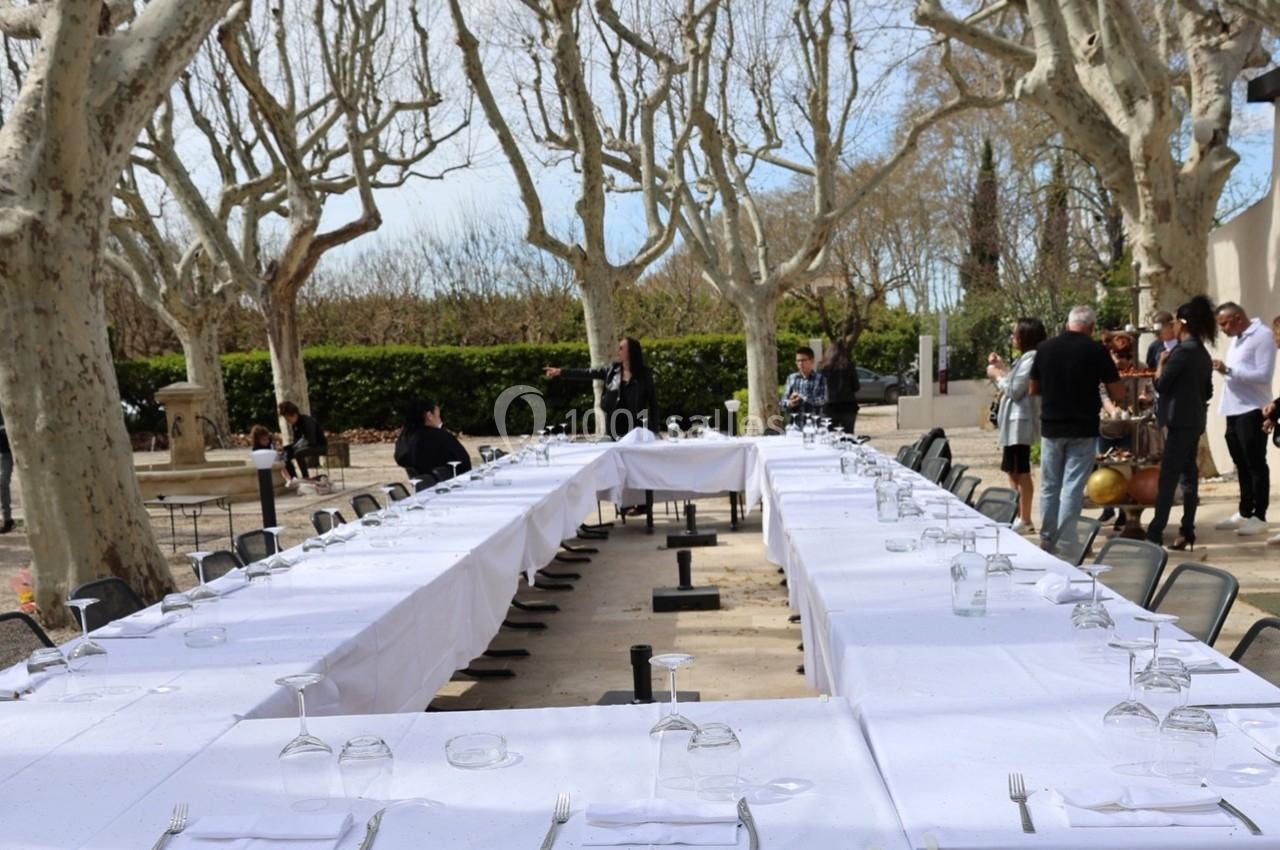 Tables dressées en extérieur sous des arbres, disposées en U avec nappes blanches, verres et couverts.