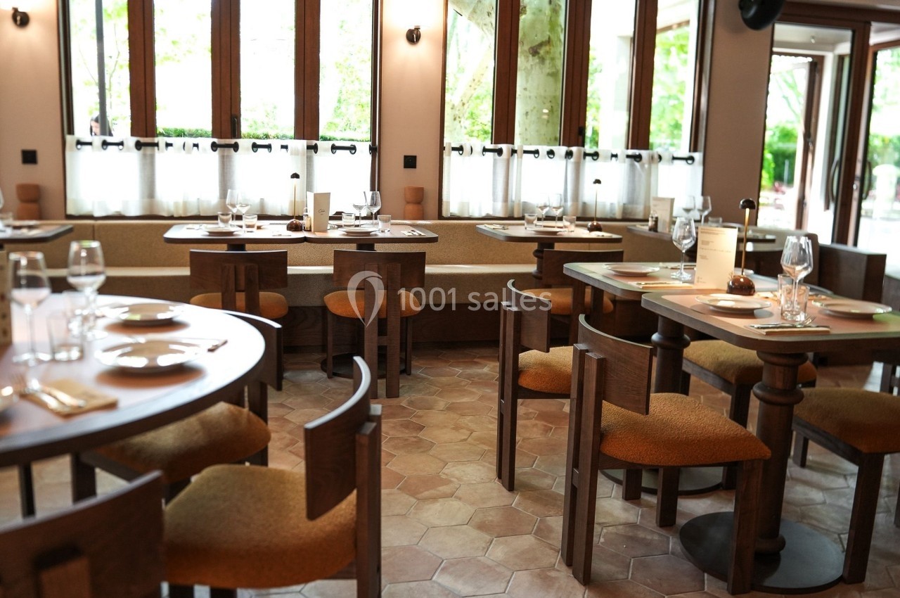 Salle de restaurant lumineuse avec tables en bois, chaises rembourrées et grandes fenêtres donnant sur l'extérieur.