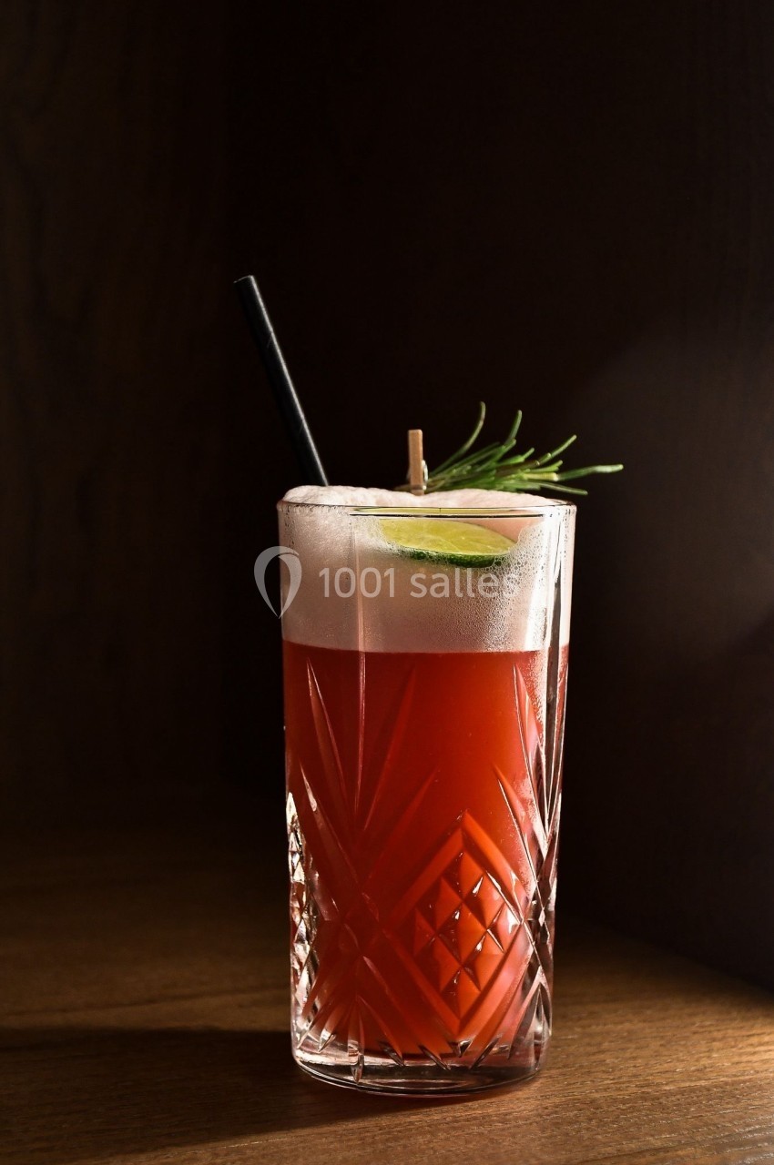 Verre à cocktail en cristal rempli d'une boisson rouge, garni d'une tranche de citron vert, d'un brin de romarin et d'une…
