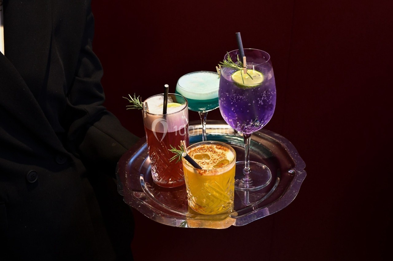 Un serveur présente un plateau contenant quatre cocktails colorés garnis de fruits et d'herbes aromatiques.