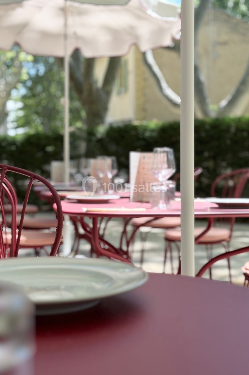 Terrasse d'un café avec des tables et chaises rouges, verres et assiettes disposés, sous des parasols blancs.