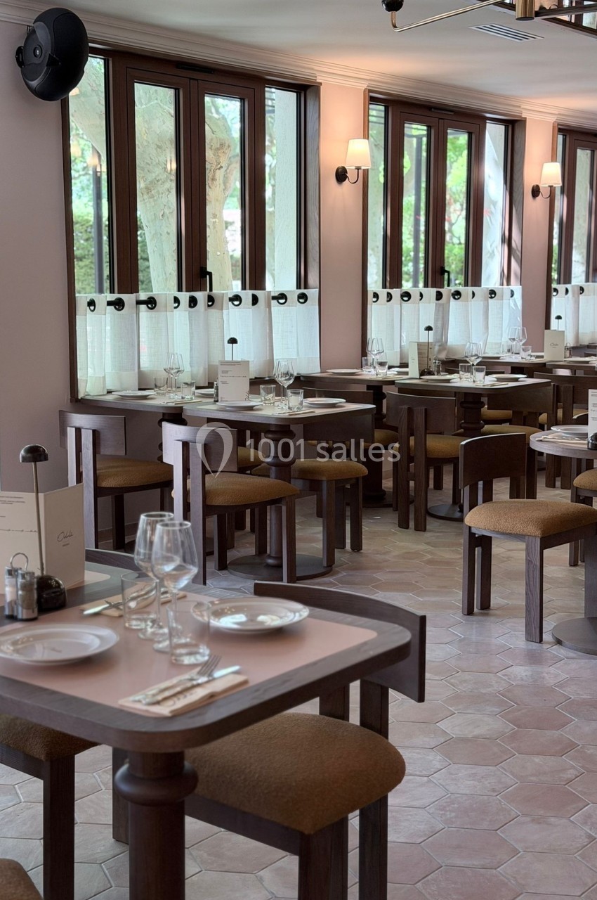 Salle de restaurant lumineuse avec tables en bois, chaises rembourrées et grandes fenêtres donnant sur des arbres.