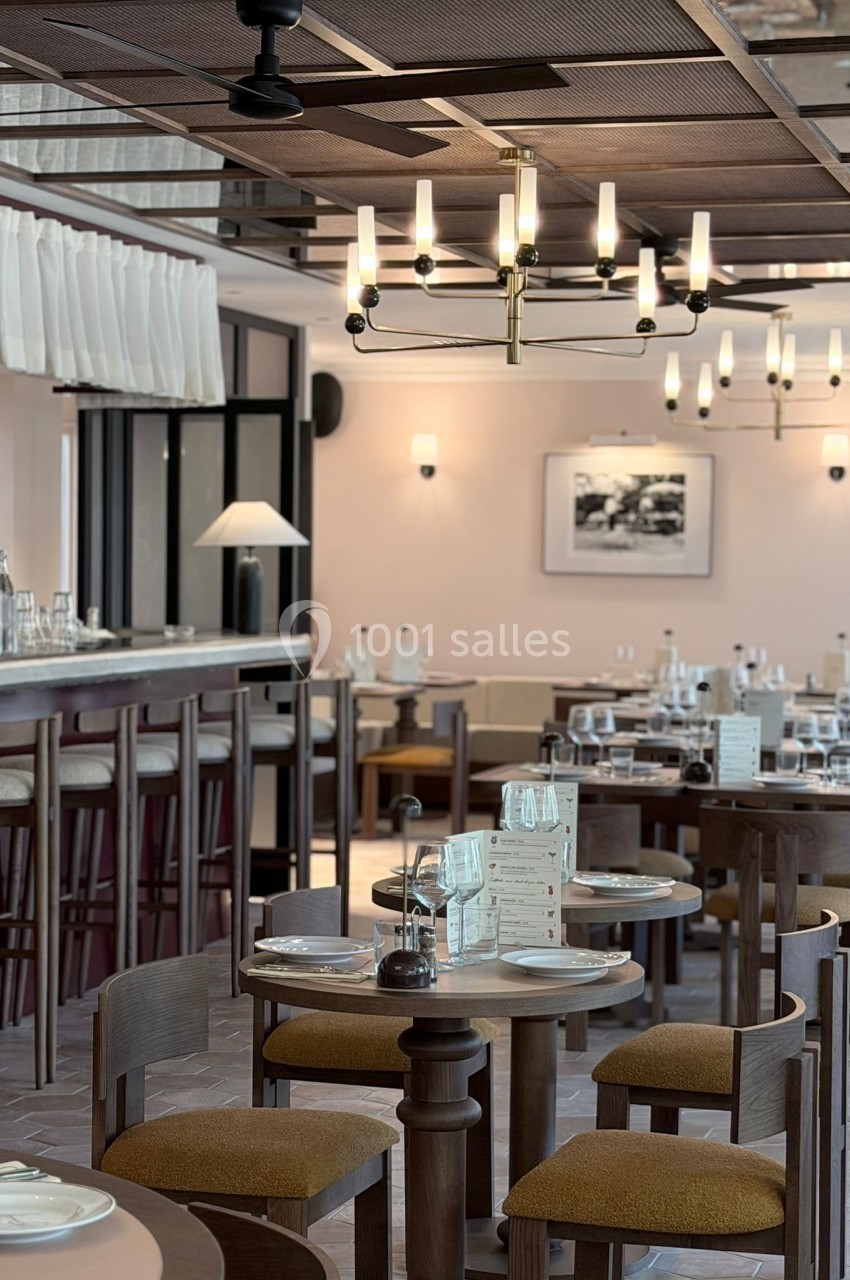 Salle de restaurant élégante avec tables en bois, chaises rembourrées, bar en arrière-plan et luminaires modernes.