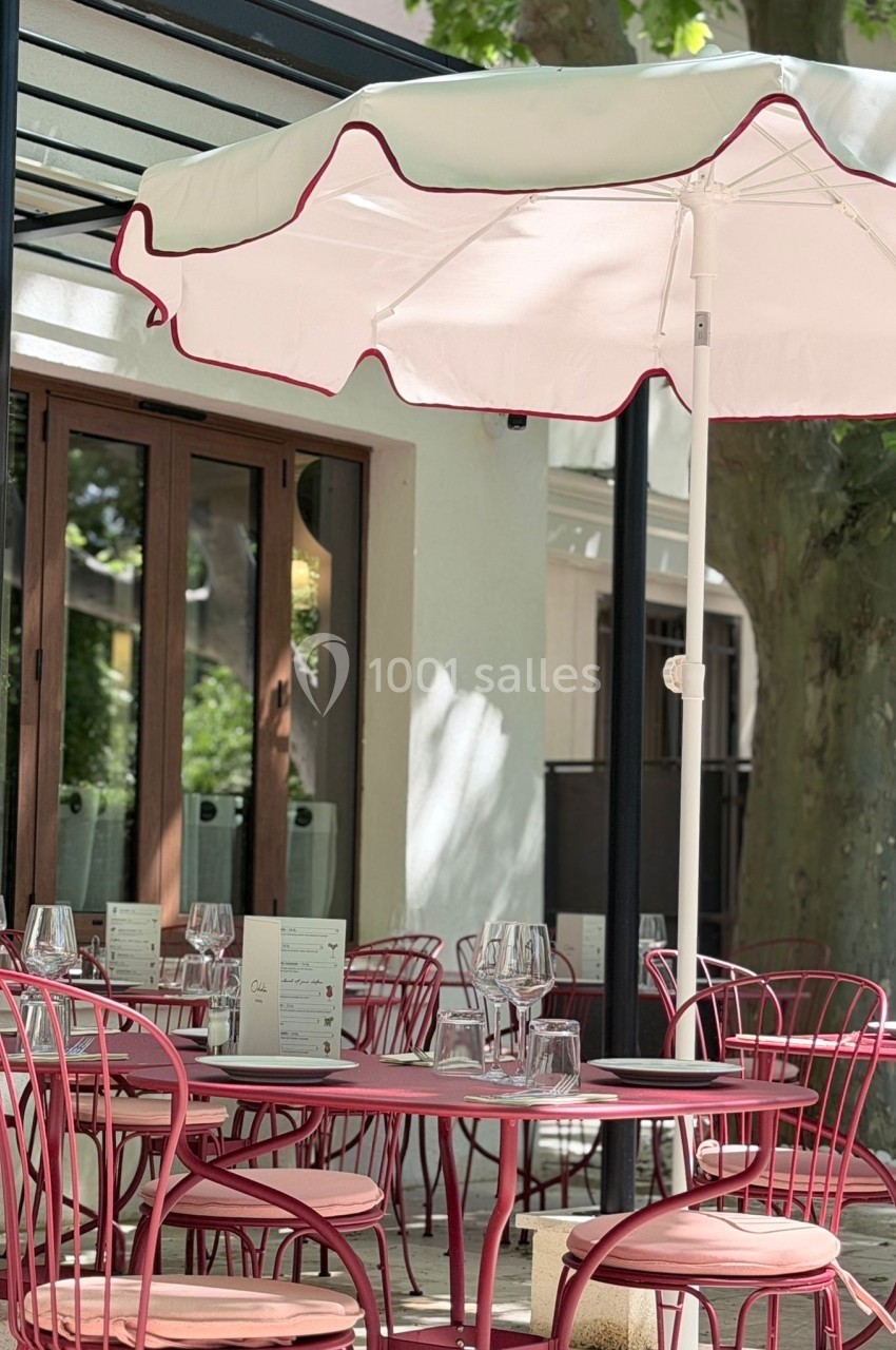 Terrasse d'un café avec des tables et chaises rouges, ombragée par un parasol blanc et entourée de verdure.