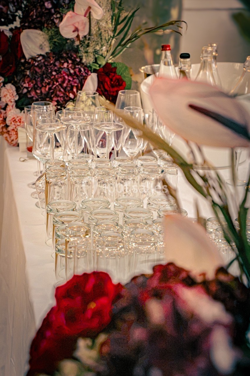Table dressée avec des verres en cristal alignés, entourée de bouquets de fleurs colorées et de bouteilles en arrière-plan.