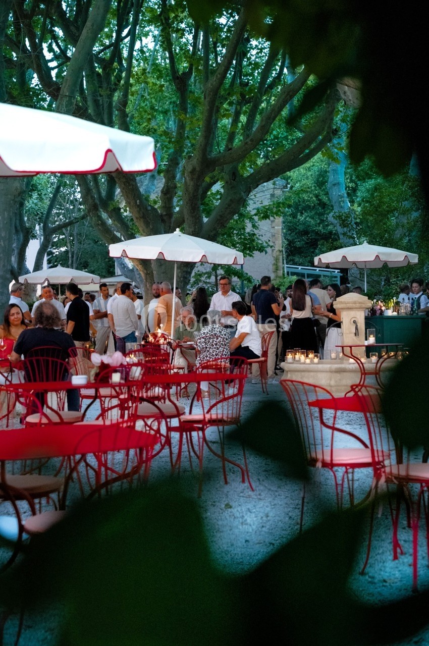 Terrasse animée en soirée avec des tables rouges, des parasols blancs et des convives sous des arbres éclairés.