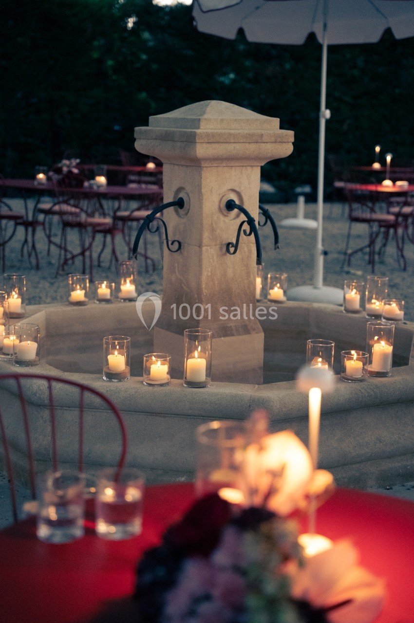 Fontaine en pierre entourée de bougies allumées dans des verres, avec des tables et chaises en arrière-plan.