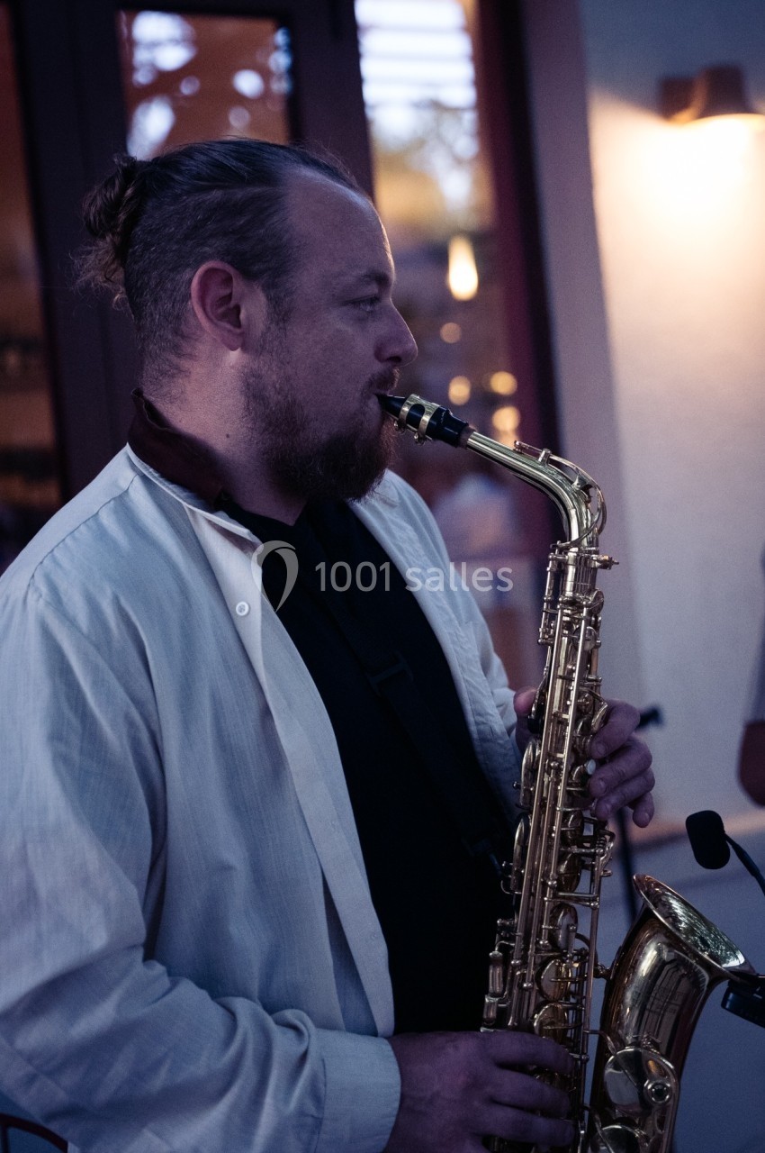 Un musicien joue du saxophone en extérieur, vêtu d'une chemise claire, devant une porte vitrée au crépuscule.