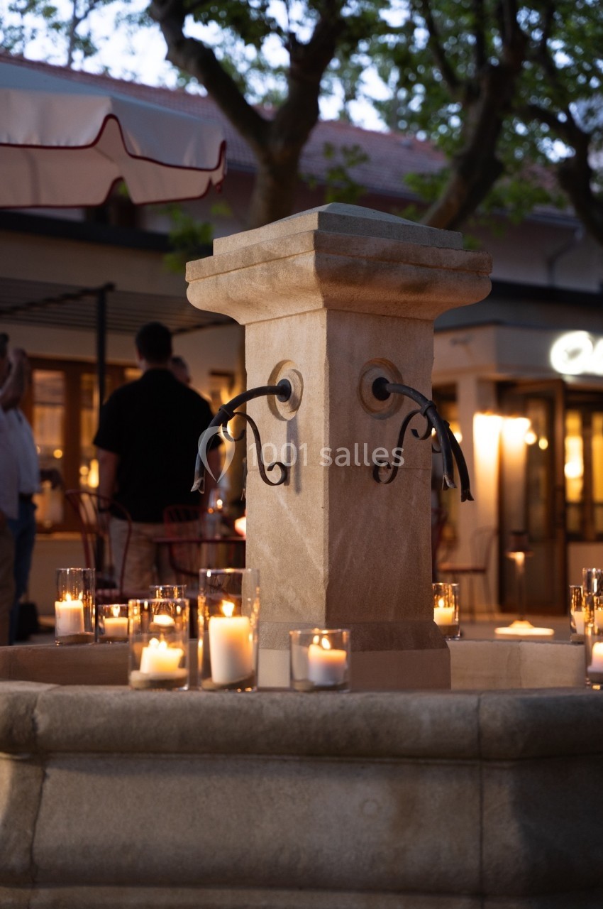 Fontaine en pierre entourée de bougies allumées, située sur une place avec des arbres et des terrasses en arrière-plan.