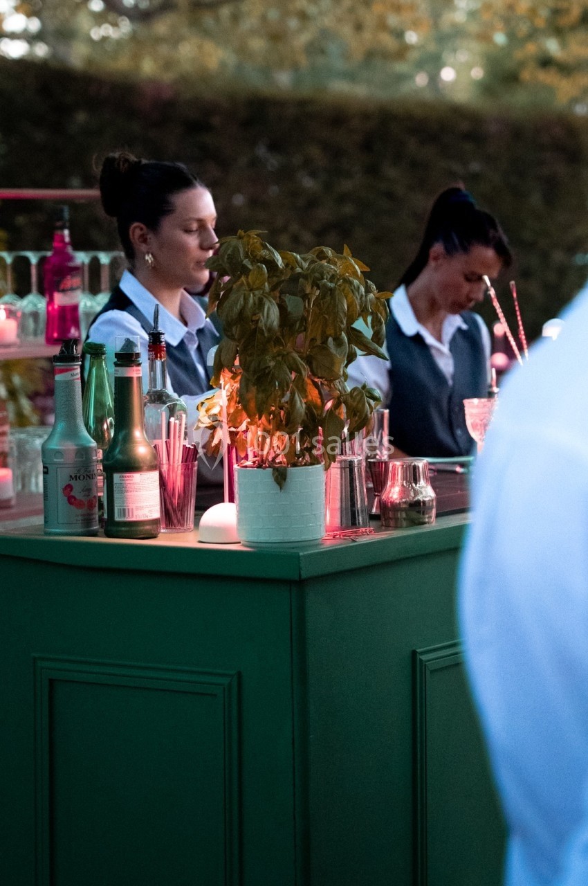 Deux serveuses derrière un bar extérieur, entourées de bouteilles et d'un pot de basilic, en ambiance tamisée.