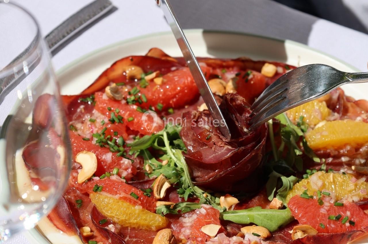 Une assiette de carpaccio de viande avec roquette, agrumes, noix et assaisonnement, une main utilise des couverts.