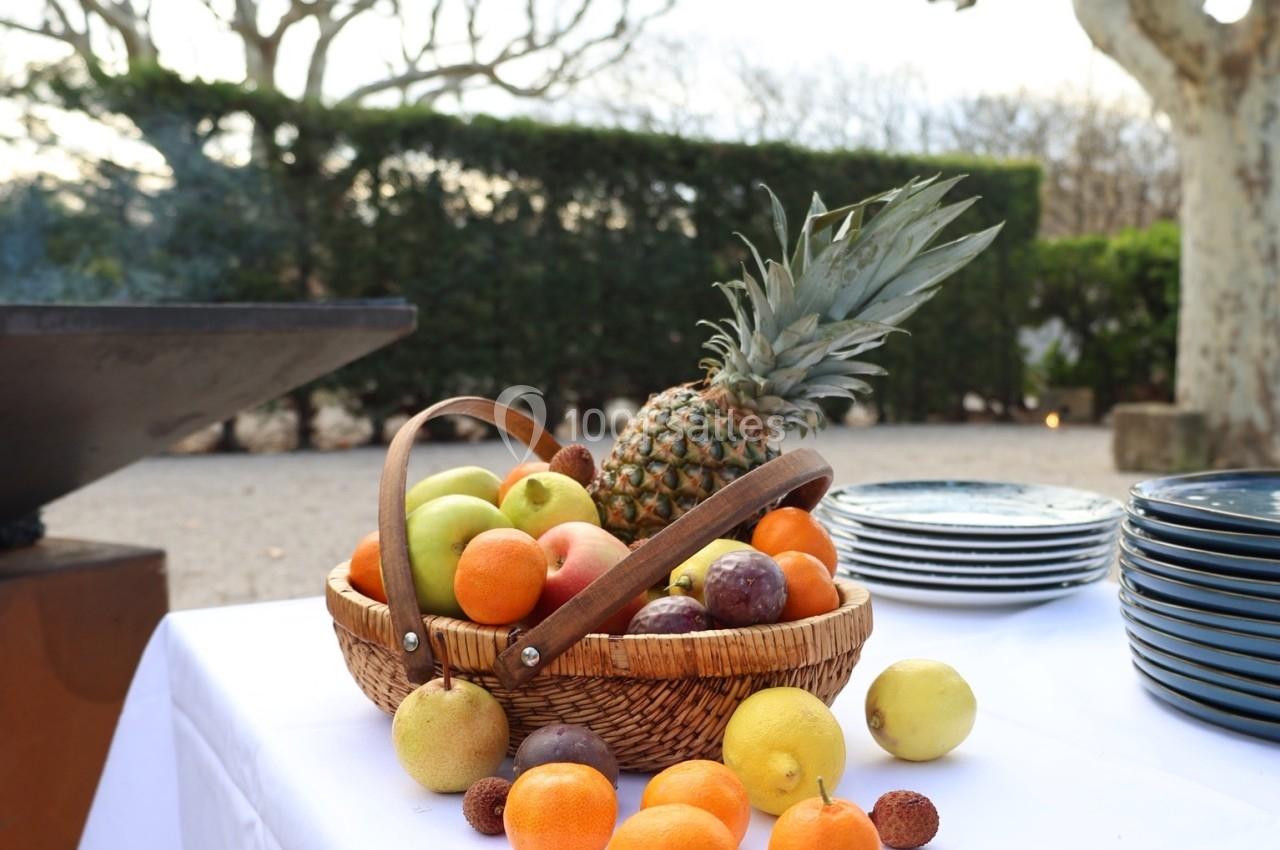 Corbeille de fruits variés sur une table blanche en extérieur, avec des assiettes empilées et des arbres en arrière-plan.