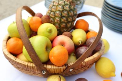 Panier en osier rempli de fruits variés, dont ananas, pommes, oranges et citrons, posé sur une nappe blanche.