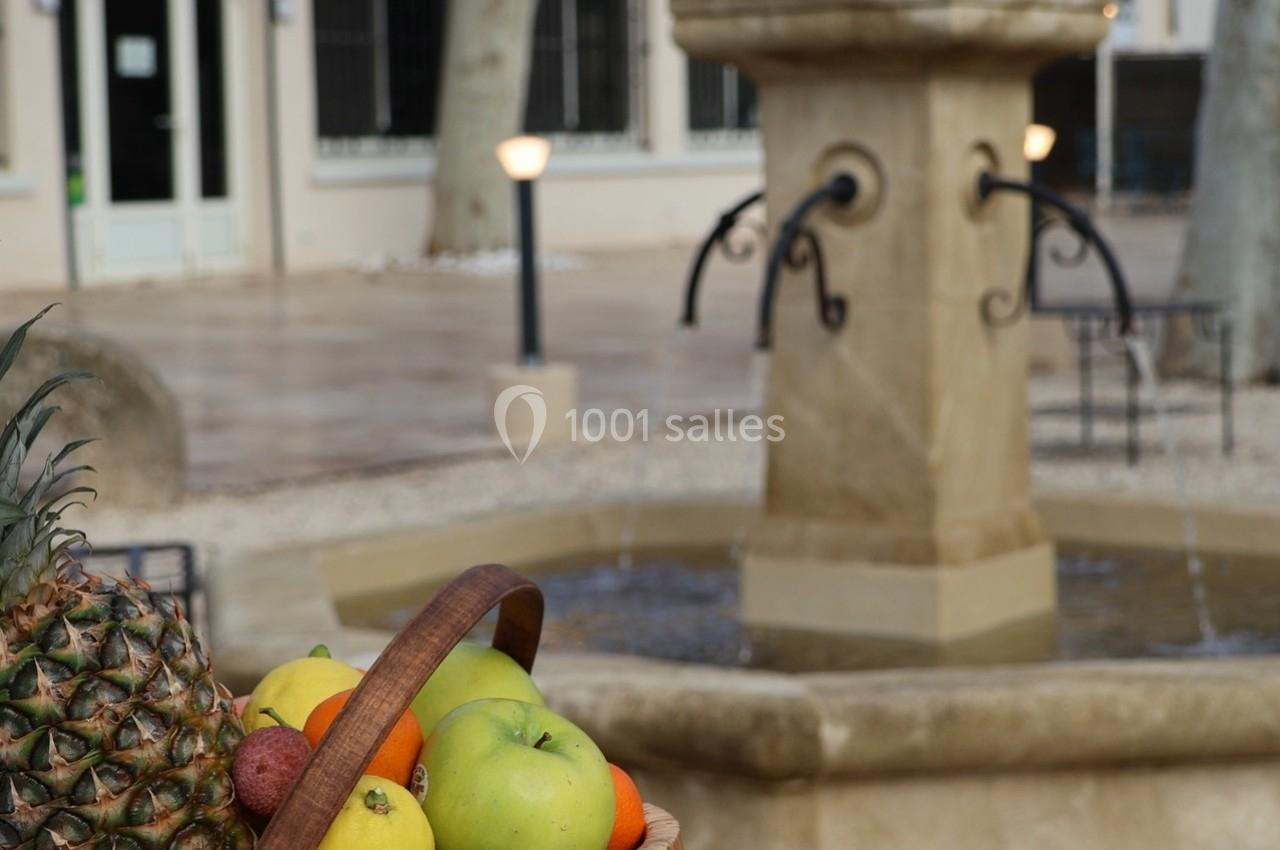 Panier de fruits variés au premier plan devant une fontaine en pierre dans une cour pavée.