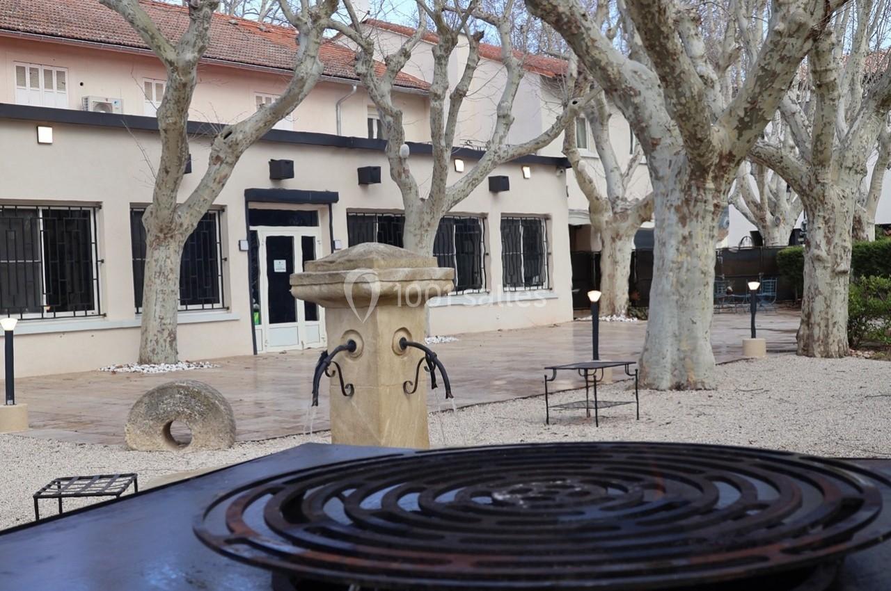 Cour extérieure avec arbres, fontaine en pierre, mobilier de jardin et bâtiment beige aux fenêtres grillagées en arrière…