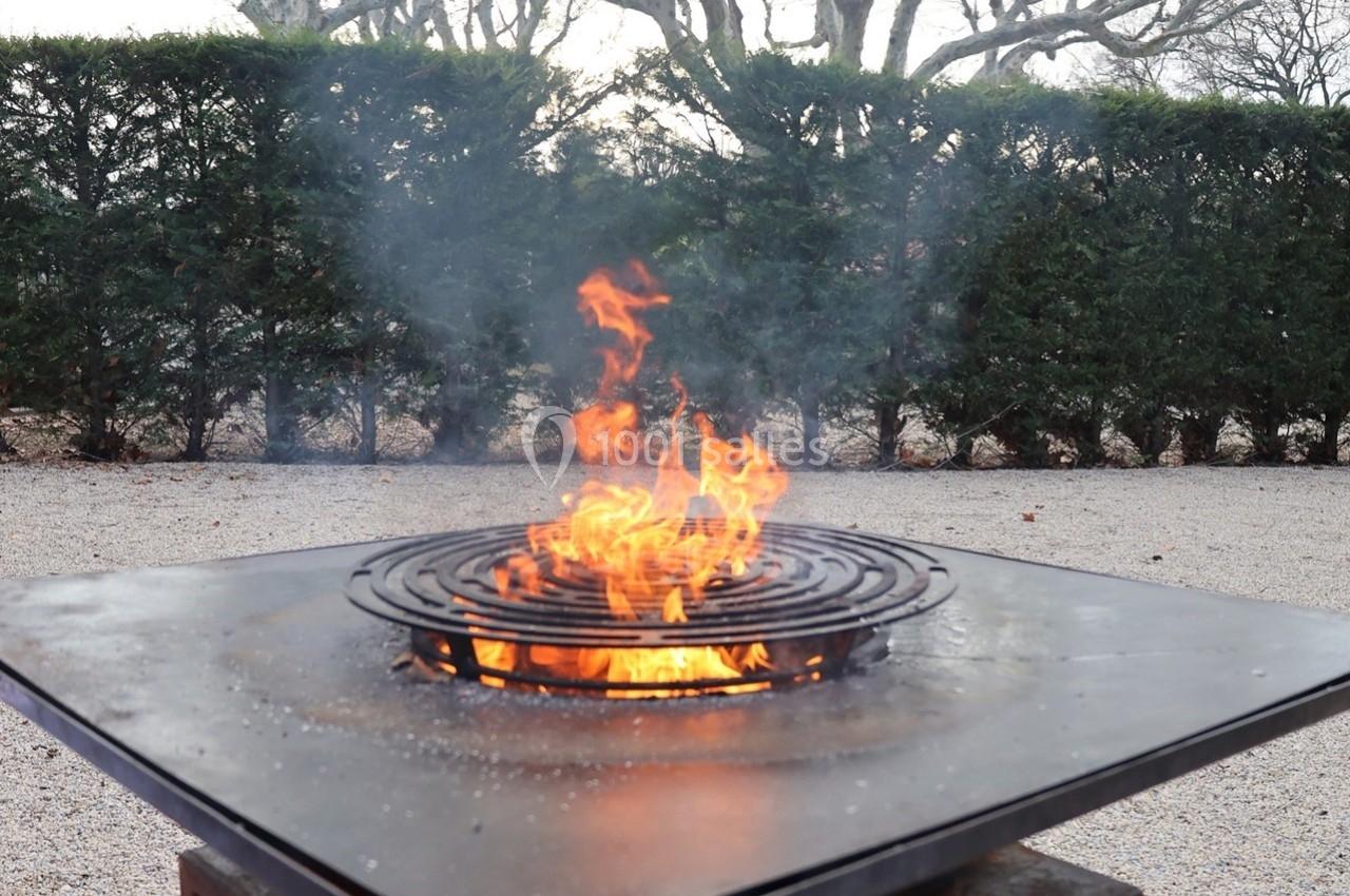 Brasero en métal avec grille circulaire, enflammé, installé sur un sol gravillonné devant une haie d'arbustes.