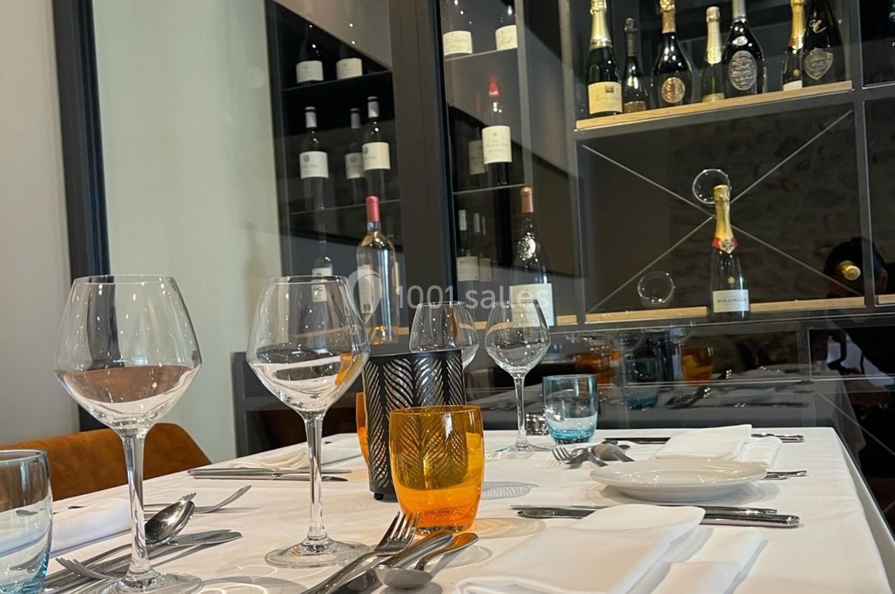 Table dressée avec verres, couverts et serviettes, devant une vitrine contenant des bouteilles de vin et de champagne.
