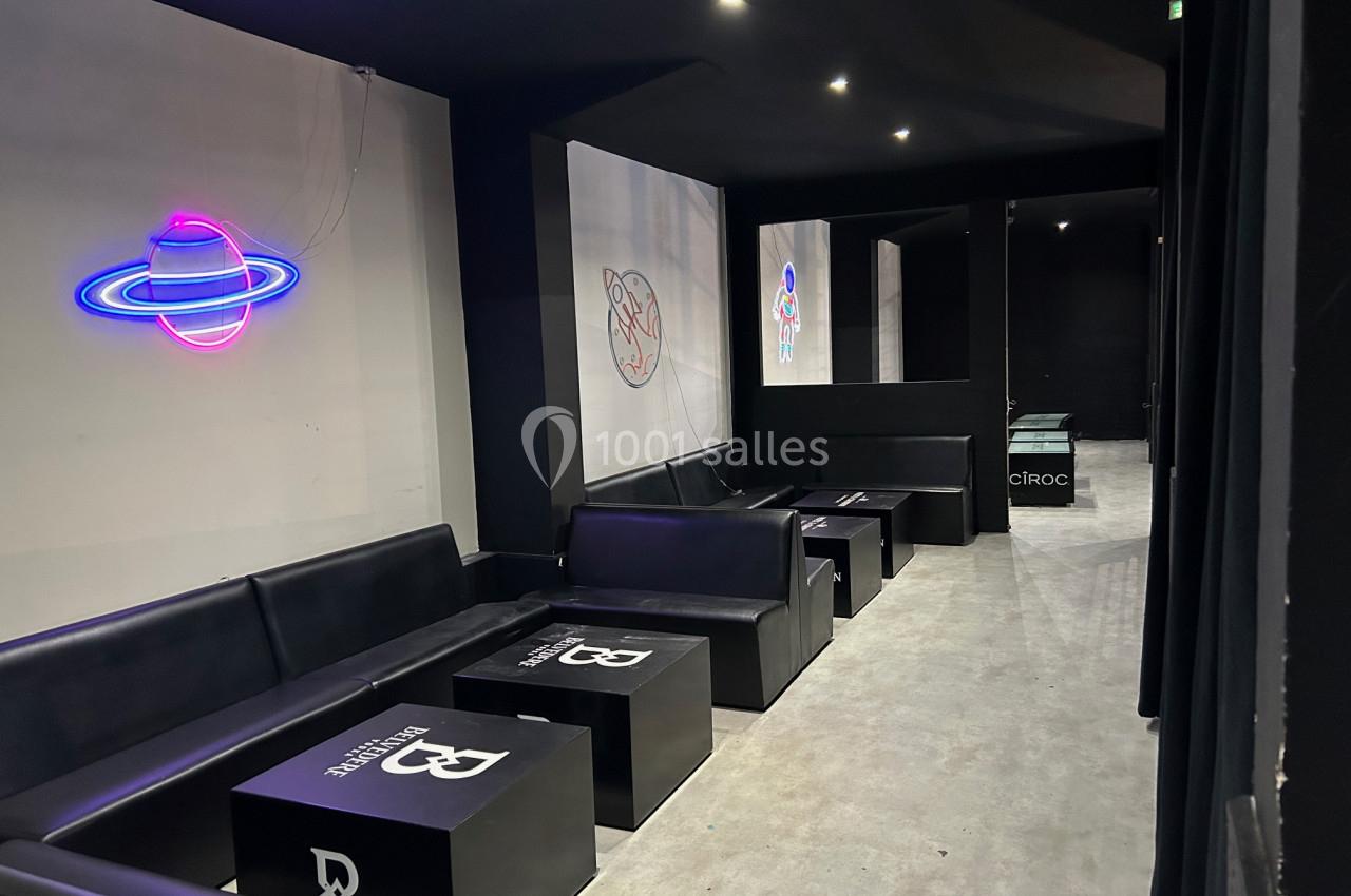 Salle moderne avec banquettes noires, tables basses et décorations lumineuses au néon sur les murs sombres.