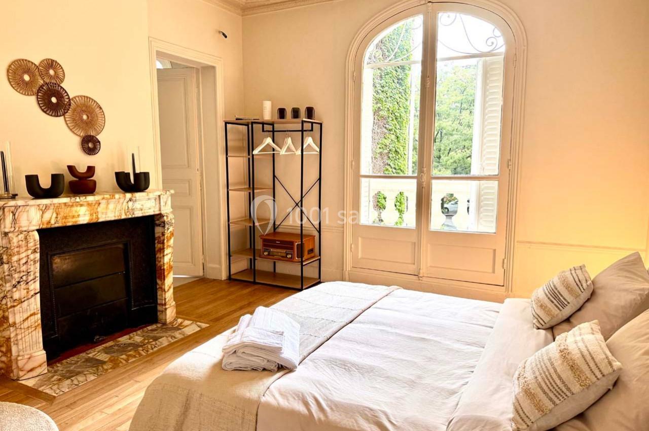 Chambre lumineuse avec lit double, cheminée décorative, étagère en métal et grande fenêtre donnant sur un jardin verdoyant.