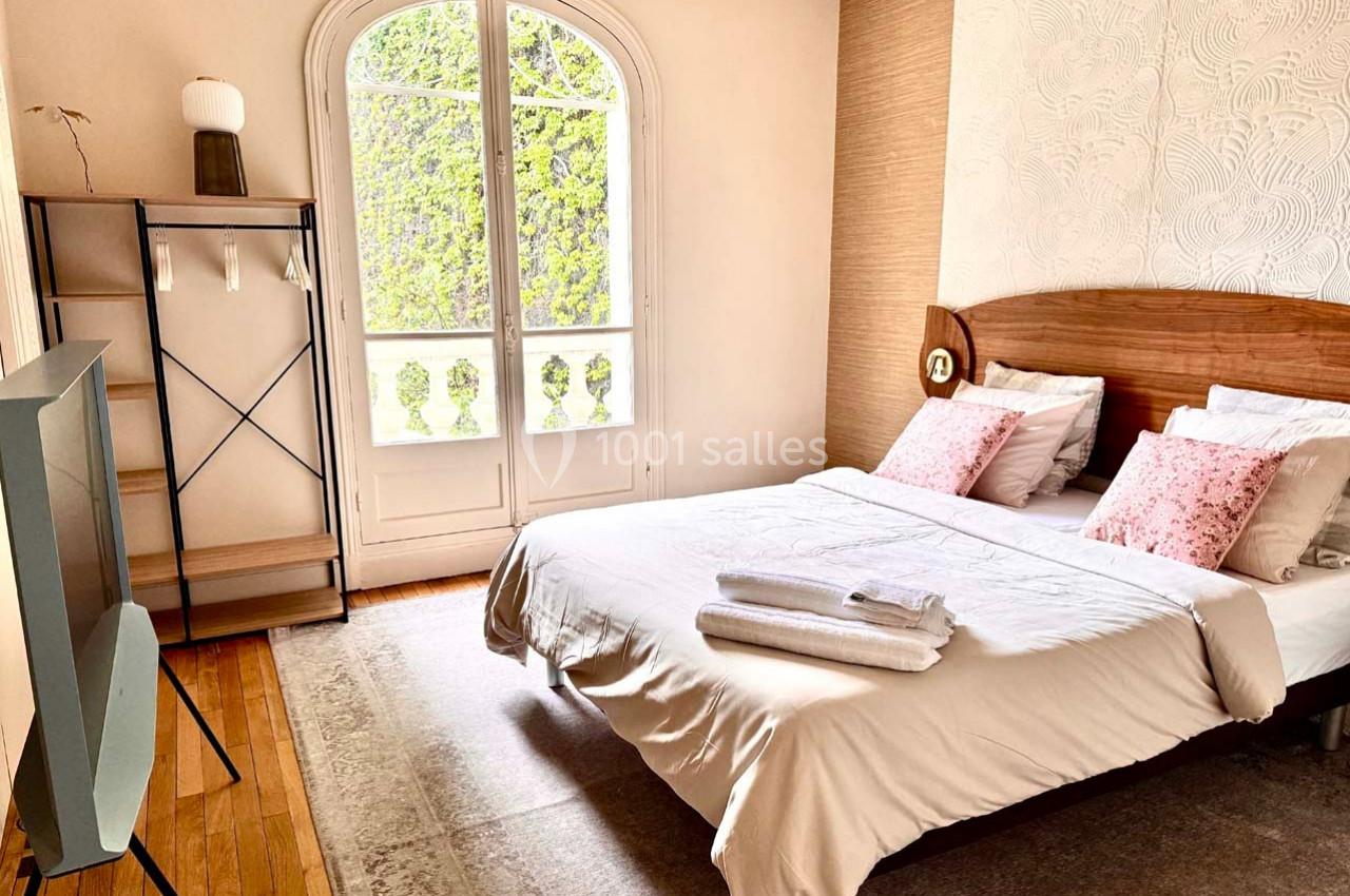 Chambre lumineuse avec lit double, étagère, télévision et grande fenêtre donnant sur des arbres.