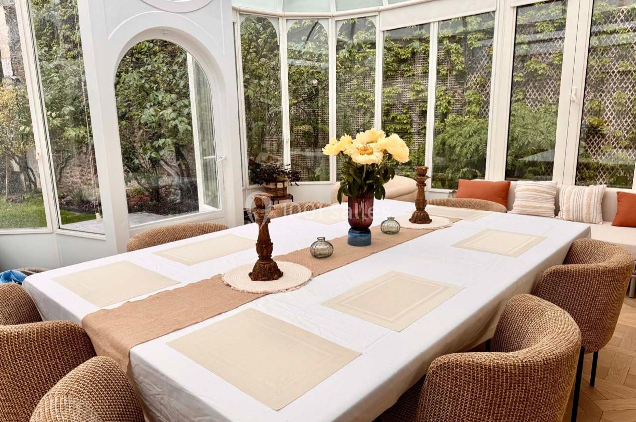 Salle à manger lumineuse avec une grande table dressée, entourée de chaises en rotin, dans une véranda avec vue sur un…