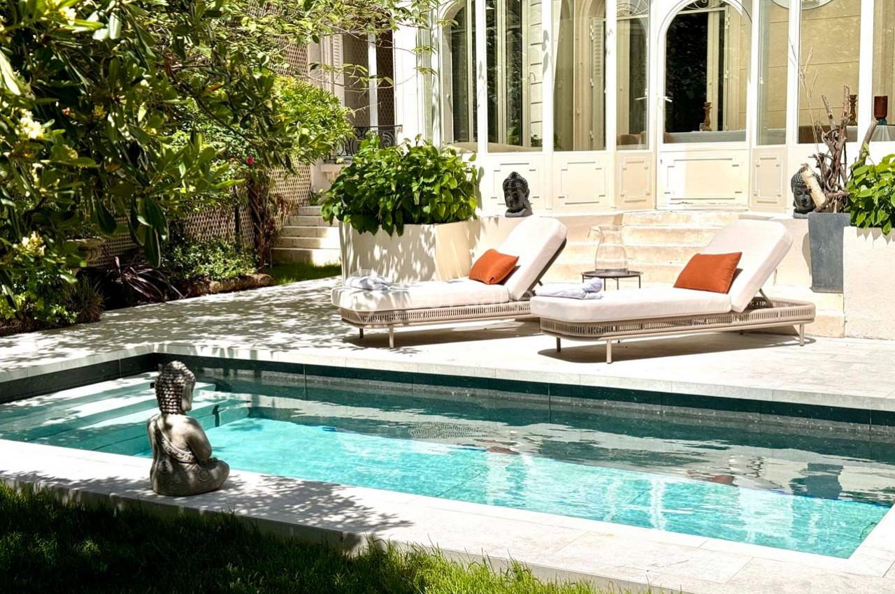 Piscine extérieure entourée de verdure, avec deux transats et une statue décorative près d'une maison lumineuse.