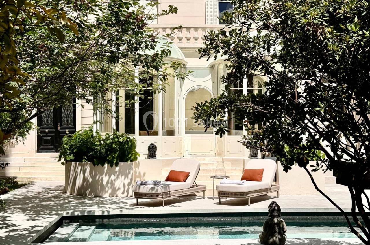 Deux chaises longues avec coussins orange près d'une piscine, entourées de végétation et devant une façade élégante.