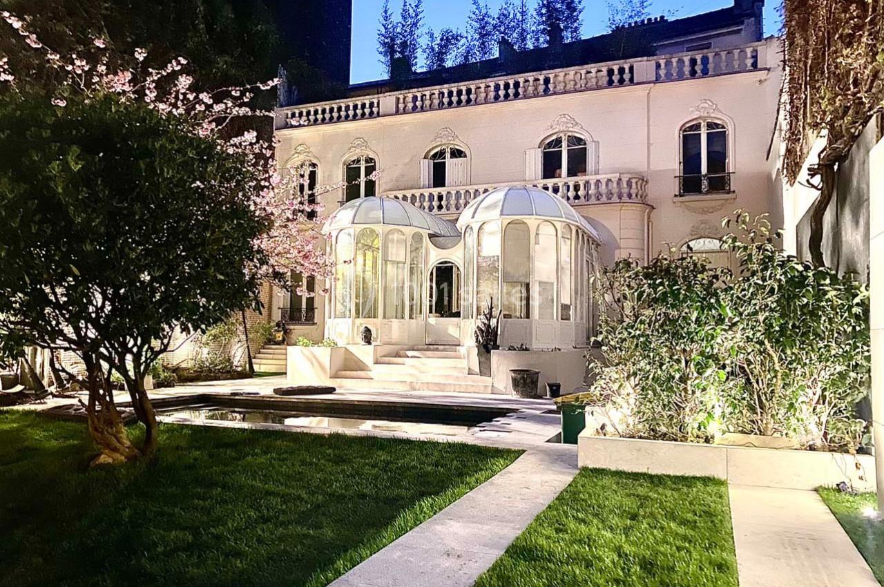 Façade d'une maison élégante avec vérandas vitrées, entourée d'un jardin éclairé et d'arbres en fleurs.