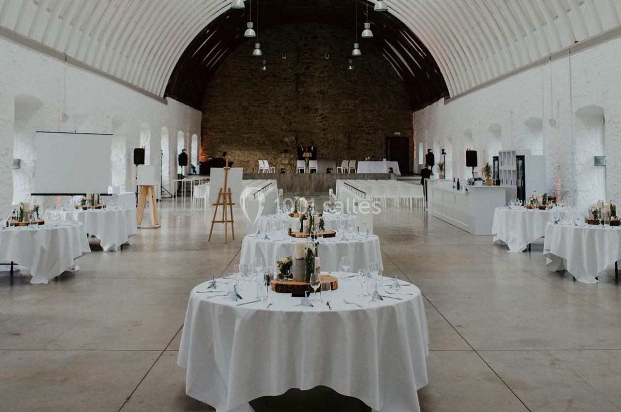 Salle de réception lumineuse avec tables rondes dressées, murs en pierre blanche et plafond voûté en bois.