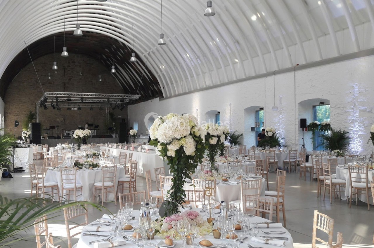Salle de réception lumineuse avec tables décorées de fleurs blanches et roses, chaises en bois clair et plafond voûté.