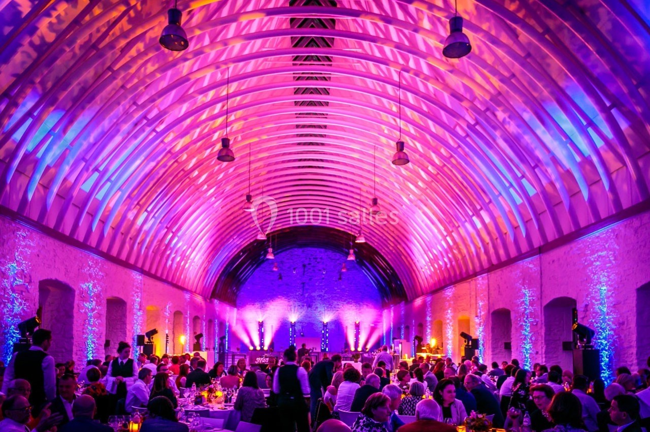 Salle voûtée illuminée par des lumières colorées, remplie de convives assis à des tables lors d'un événement.