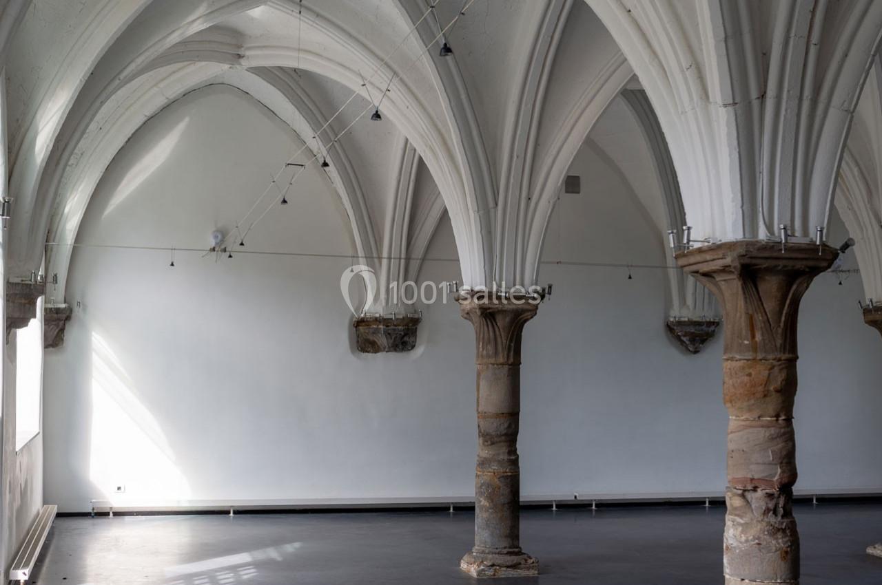 Salle vide avec des colonnes en pierre et des voûtes gothiques blanches sous un éclairage naturel.