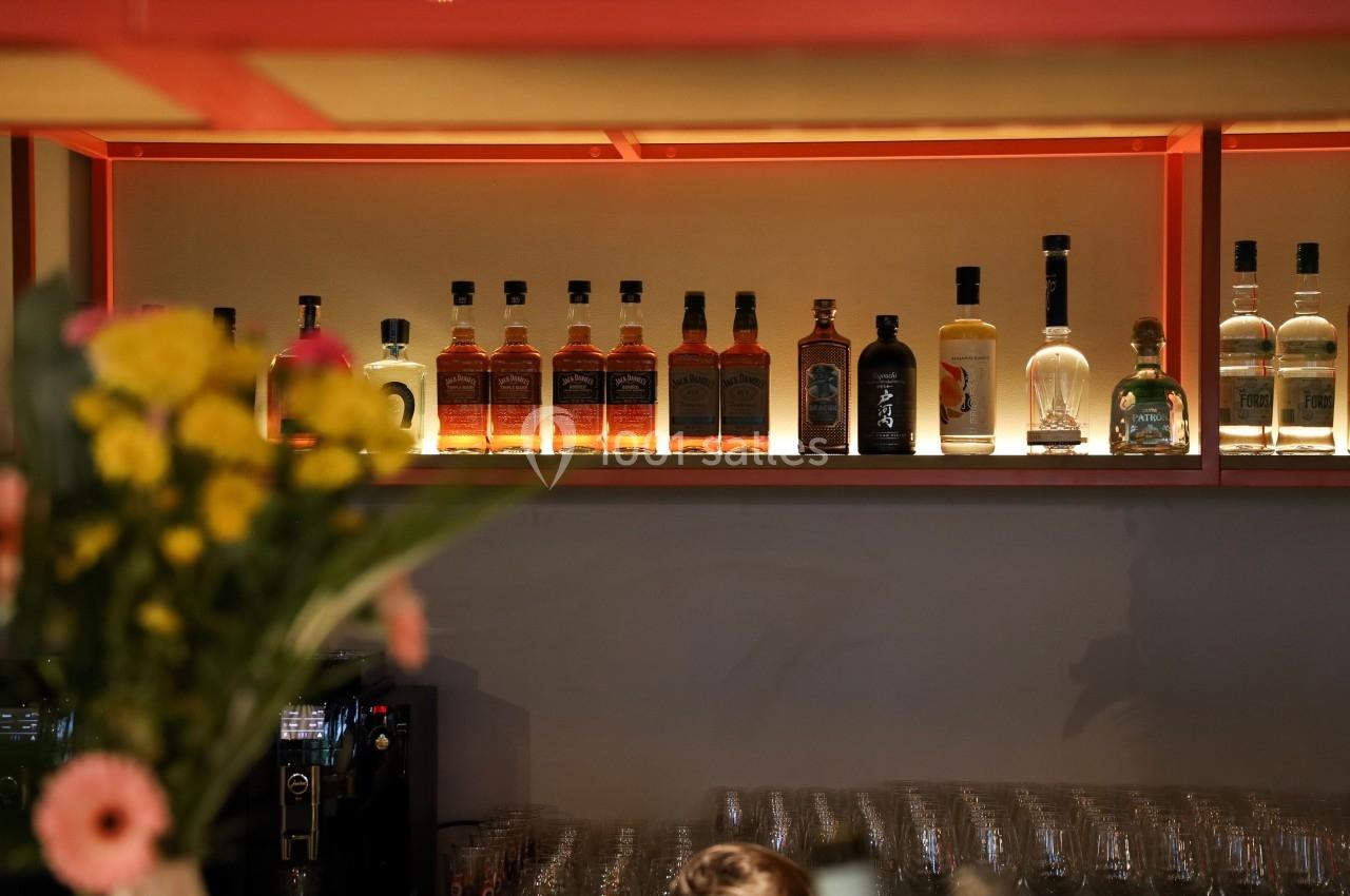 Bouteilles d'alcool alignées sur une étagère éclairée, avec un bouquet de fleurs flou au premier plan.