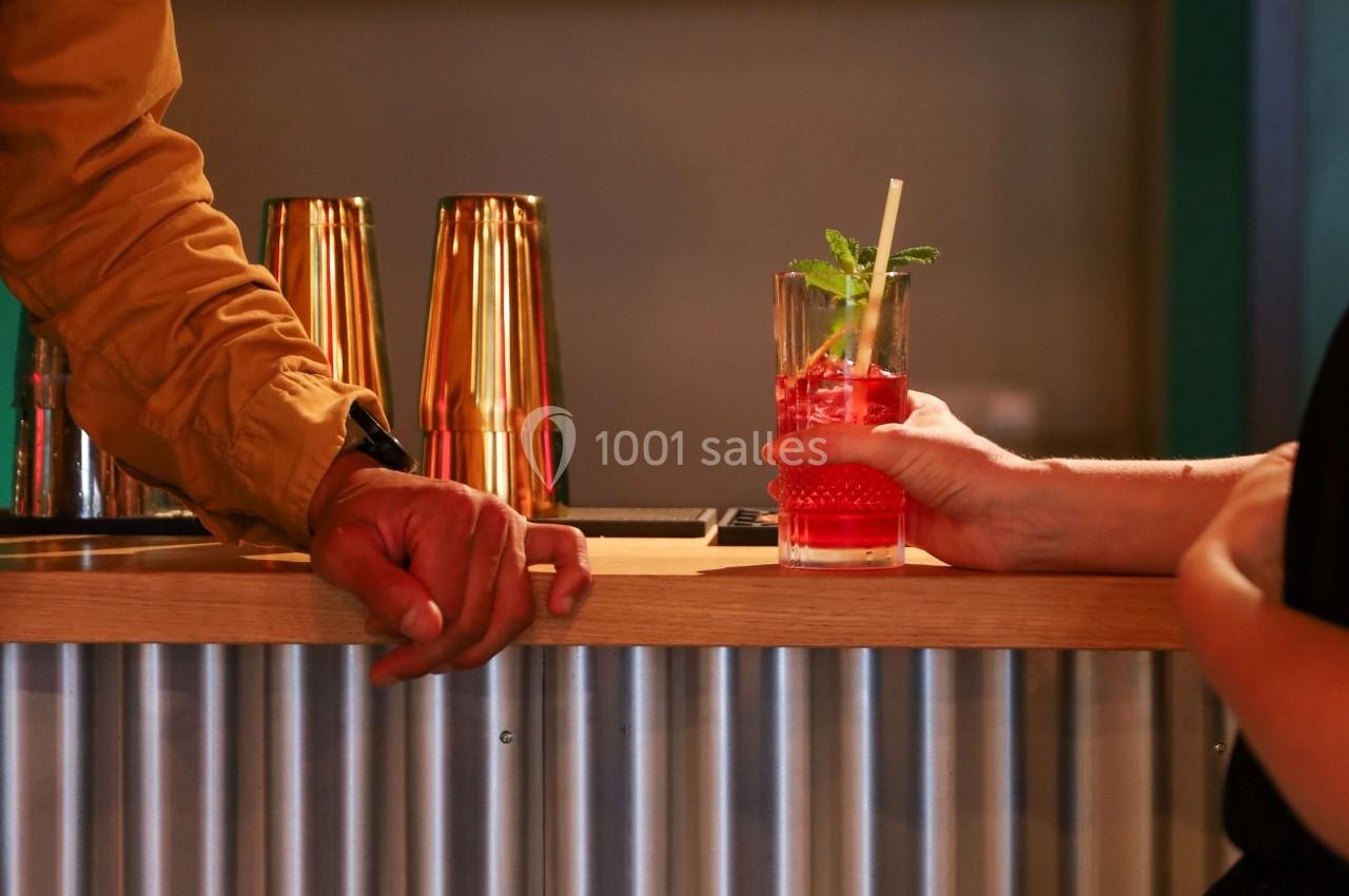 Un verre de cocktail rouge avec une paille et une feuille de menthe est tendu sur un comptoir de bar en métal.