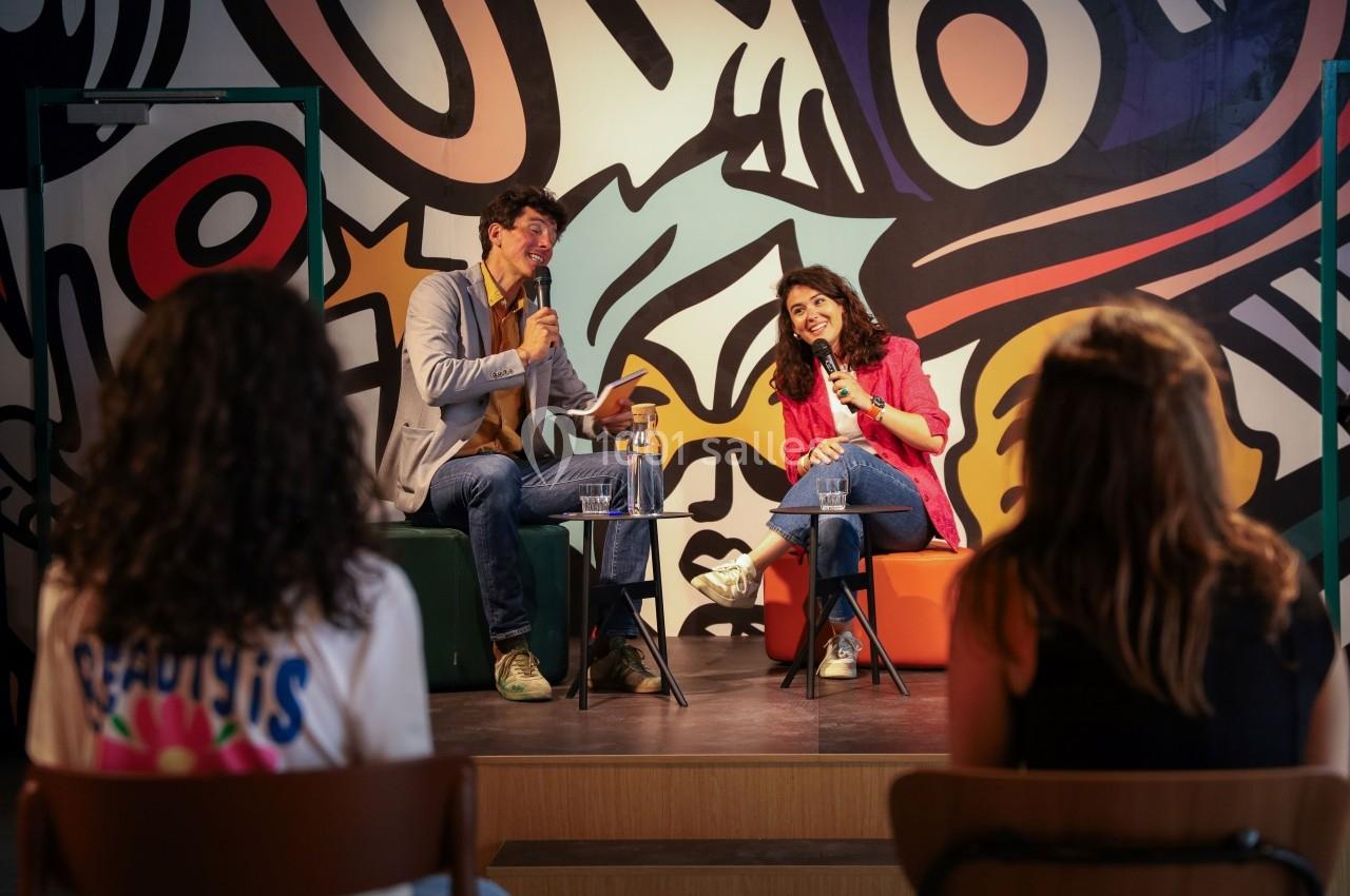 Deux personnes discutent avec des micros sur une scène devant un public, devant une fresque colorée.