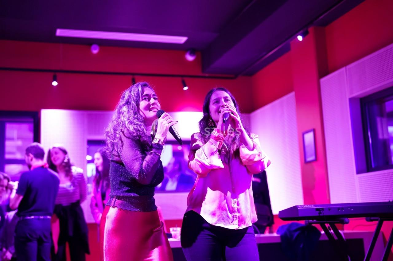 Deux femmes chantent au micro dans une salle éclairée de lumières violettes, avec des spectateurs en arrière-plan.