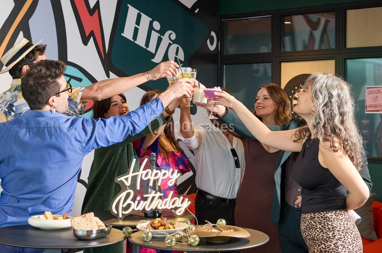 Un groupe de personnes trinque autour de tables hautes lors d'une fête d'anniversaire dans un espace intérieur décoré.