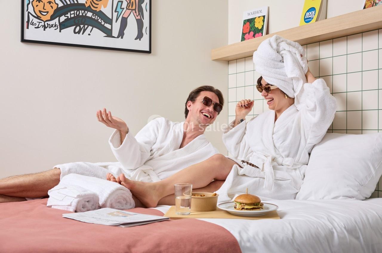 Deux personnes en peignoirs discutent et rient sur un lit, entourées de nourriture et d'objets de détente.