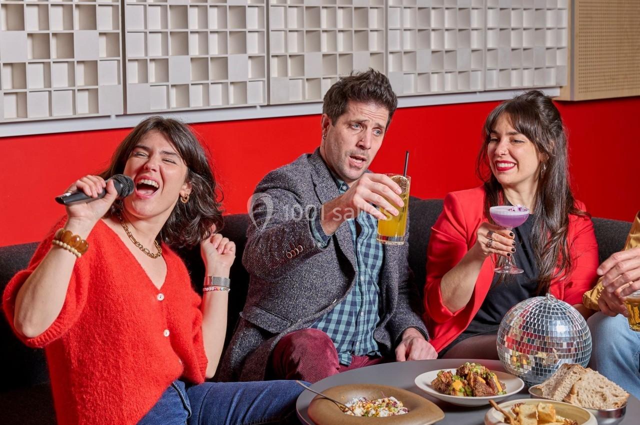 Trois personnes assises dans un salon rouge, partageant des boissons et chantant avec un micro, ambiance conviviale.