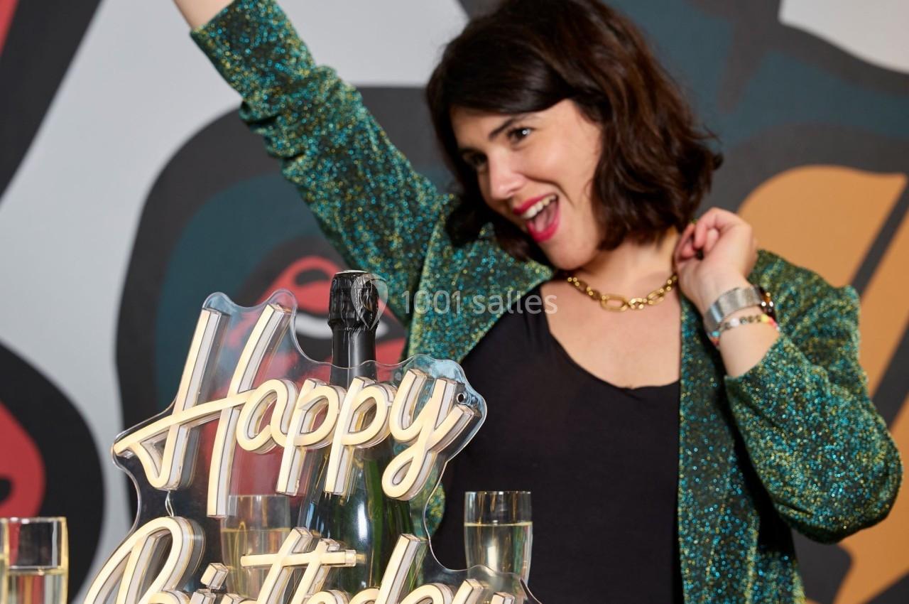 Femme souriante en tenue festive levant le bras près d'une table avec une décoration ’Happy Birthday’ et des verres.