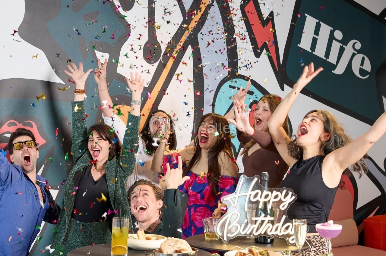 Un groupe de personnes célèbrent un anniversaire avec des confettis, des boissons et un gâteau dans une ambiance festive.