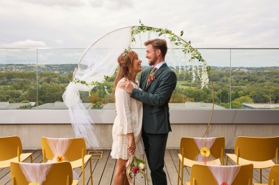 Un couple célèbre son mariage sur une petite scène décorée de voiles et de feuillages, devant une fresque colorée.